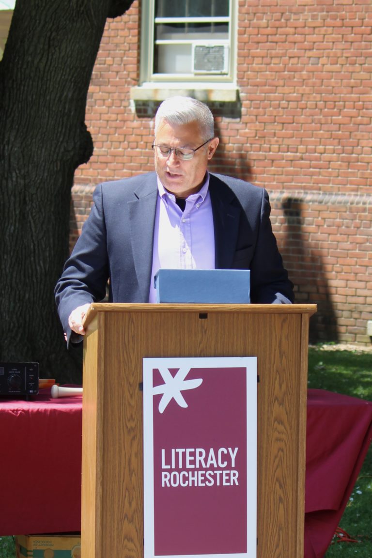 John Roman at podium with Literacy Rochester logo