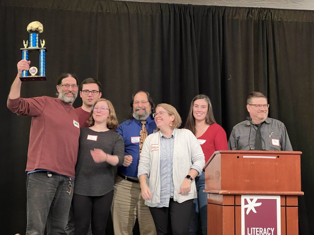 Brain Game 2022 Champions: Monroe County Library System (Seven people standing on stage, one holding trophy in the air)